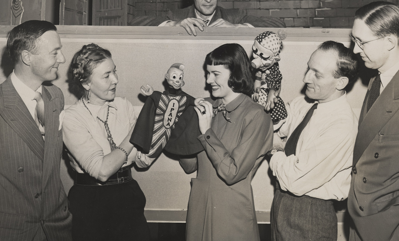 Five smiling people stand in a row, the third and fourth holding puppets