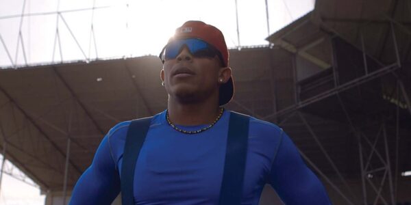 A man standing with sunglasses, a red backwards cap, and a tight blue shirt looking past camera