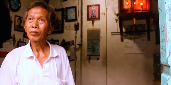 An older man in a white shirt looks out of frame as he stands in a beige room.