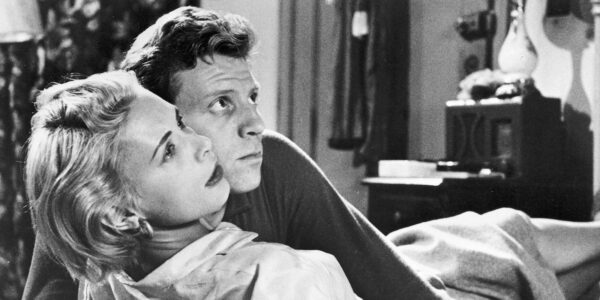 A man embracing a woman in bed; both are tense, looking to the left out of frame