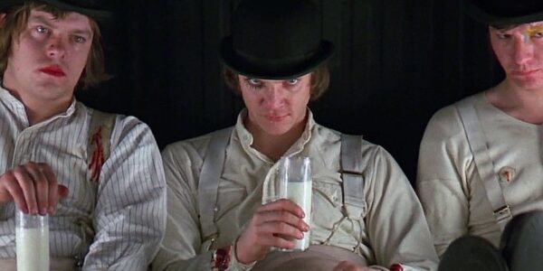 A sneering man in a bowler hat, holds a glass of milk and looks straight at the camera