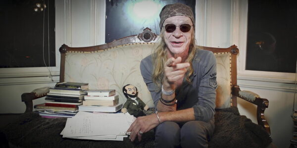A man with long hair and in sunglasses sits on a couch and points at the camera