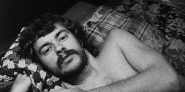 A bearded, shirtless man reclines in bed and looks at camera.