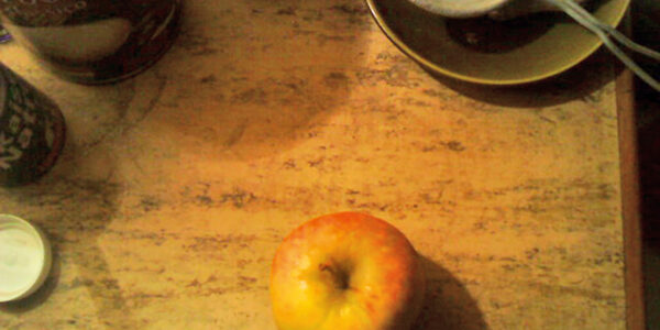 A photographic still life of an apple on a table, with mug and saucer nearby.