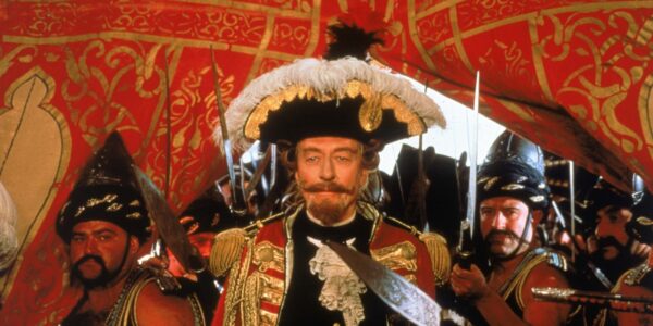 A man in a goatee and Napoleon hat underneath a deep red tent.
