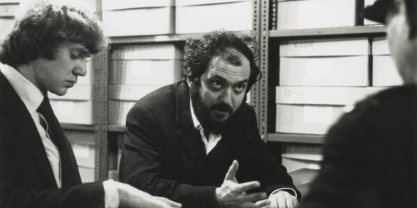 Stanley Kubrick sits at a table and looks off camera, sitting next to Malcolm McDowell