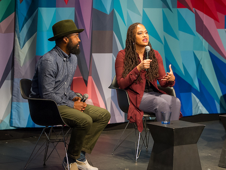 A man in a hat and a woman in a maroon jacket—cinematographer Bradford Young and director Ava DuVernay—speak to each other on-stage at Museum of the Moving Image, before a brightly colored kaleidoscopic curtain.