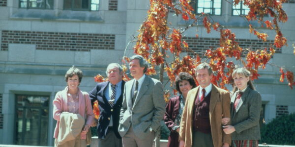 A group of six adult friends line up in front of a brightly colored autumn tree.