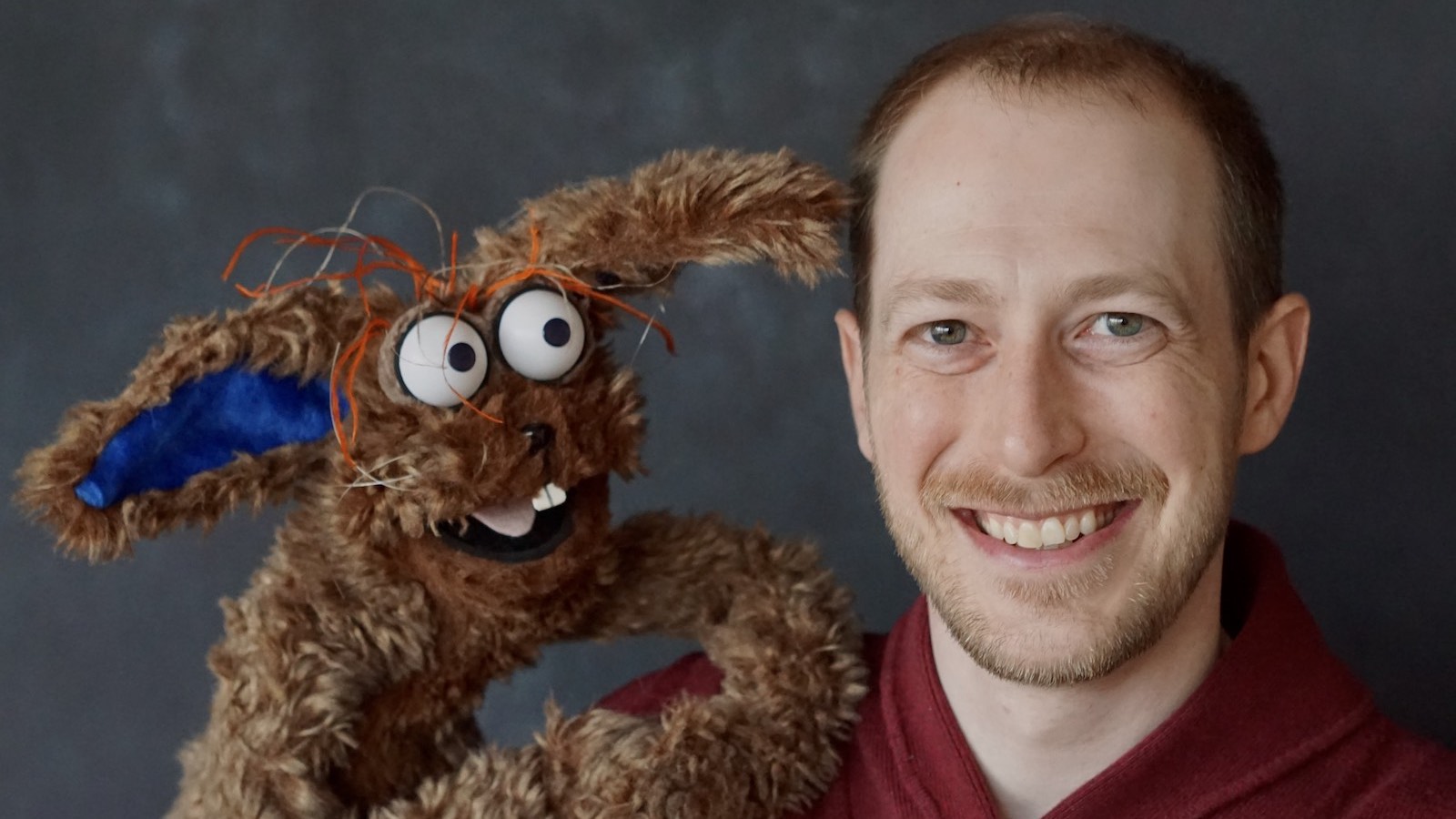 Carson SillyWilly LG Puppeteer Brian Carson smiles at camera in closeup next to his puppet, the brown rabbit Silly Willy looking at him