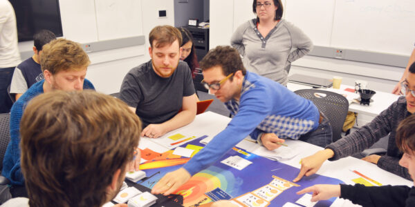 A group of people play a Role-Playing game; a man in glasses wearing a blue shirt reaches across the table