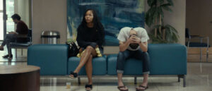 A woman and man sit next to each other in a waiting room; she looks up, his head is buried in his hands.