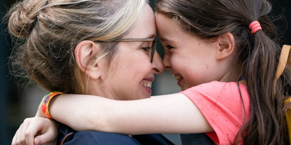 A blonde mother hugging her ponytailed daughter and both smiling