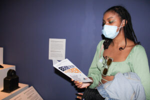 A masked young woman in the Museum galleries holds a piece of museum brochure paper and reads it.
