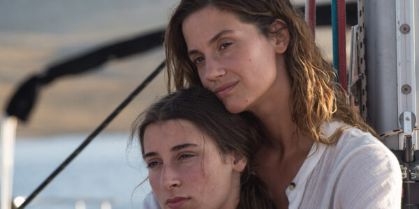 A middle aged woman in white holds a young woman, her daughter from behind in a warm embrace,