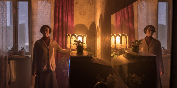 A woman stands by a lamp and a plant atop a glowing television screen in a curtained room