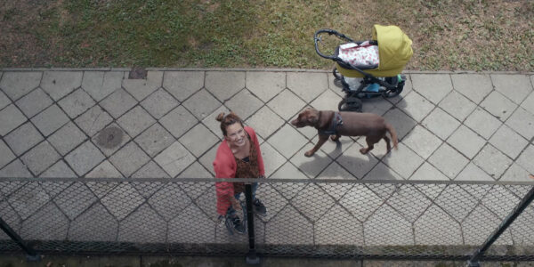 A high-angle shot of a smiling woman looking up at a balcony, with her dog and a baby carriage close by