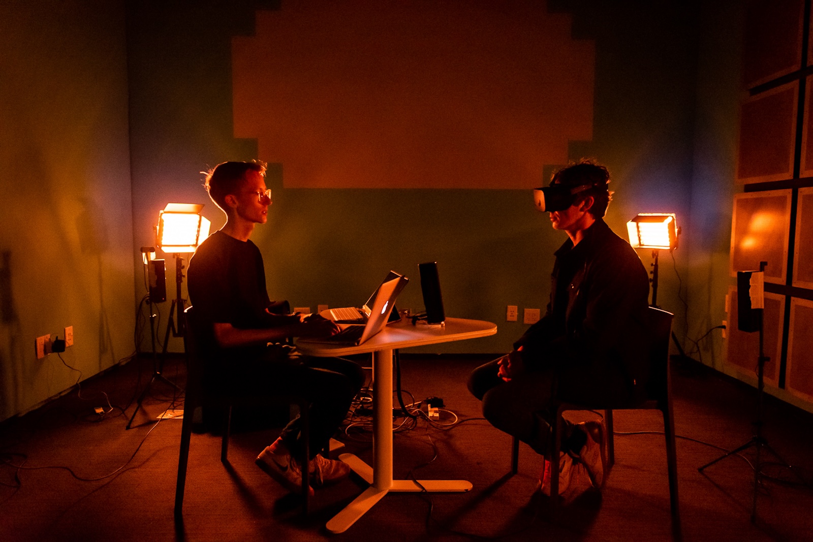 In this photograph, two seated figures face each other acrorss a triangular white table. The one on the left wears glasses and has an open laptop; the one on the right has dark hair and is wearing a VR headset. The smallish room is tinged in reddish-orange light emanating from a set of two floodlamps on each side of the people.