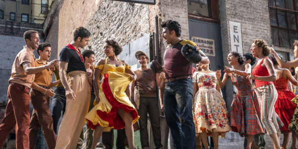 A dancing woman in bright yellow and red-trimmed dress stands between two men on a sidewalk