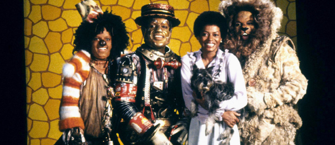Four cast members of The Wiz, including Dorothy holding her little dog Toto, look at the camera and smile.