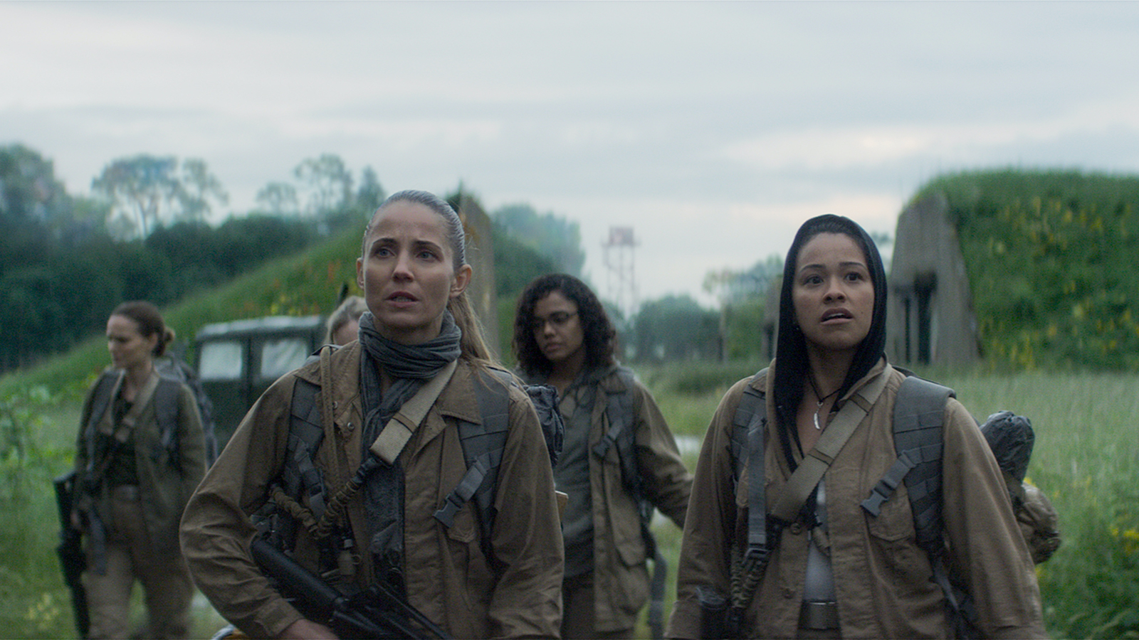 Five armed women in rugged outdoor gear scout a grassy location