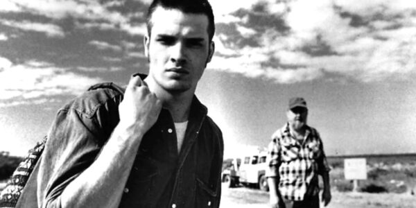 A black and white image of a young man looking into camera on an open road, with clouds in the sky overhead