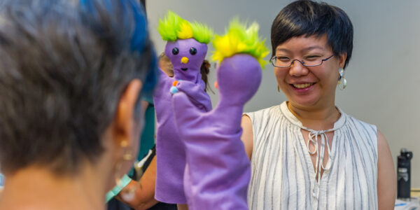 Two women smile and hold purple puppets up to each other