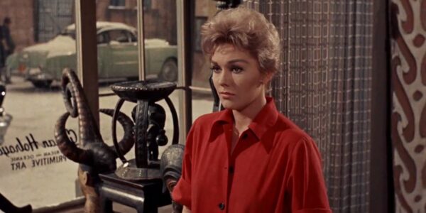 A blonde woman in a red button down shirt looks off-screen while standing in front of a window