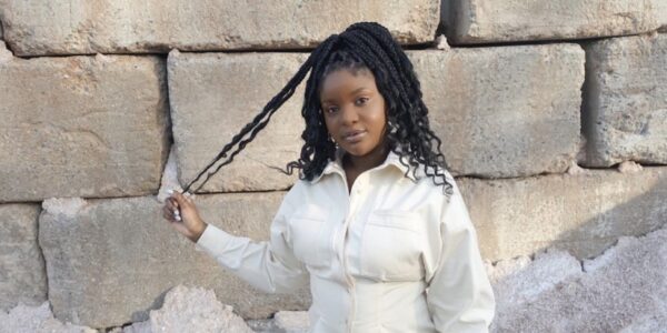 A young woman with long braided hair wears a whilte long-sleeved button-down blouse and stands in front of a wall with large stone blocks