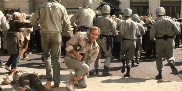 A man crouches on the ground with a gun, surrounded by men in uniform