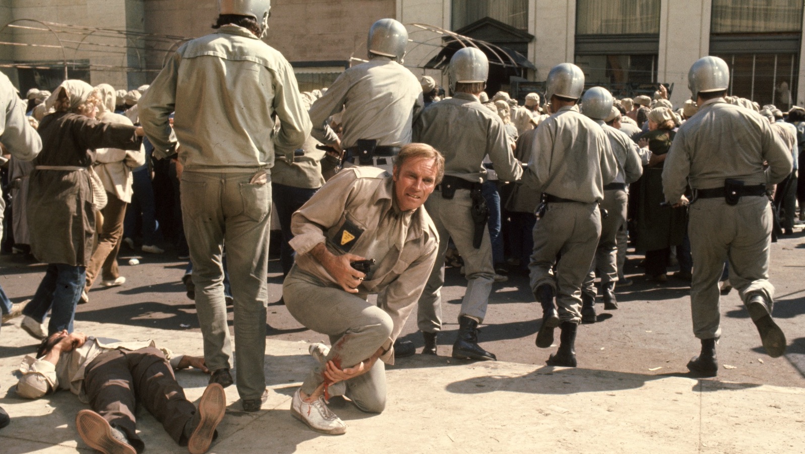 A man crouches on the ground with a gun, surrounded by men in uniform