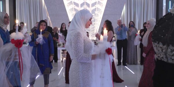 A woman in a white dress and hijab in profile holds a candle in a long hallway with others walking past her