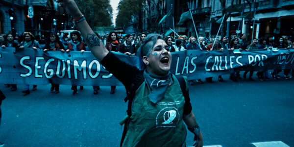 A woman marches in the street in front of a banner, one arm raised in triumph