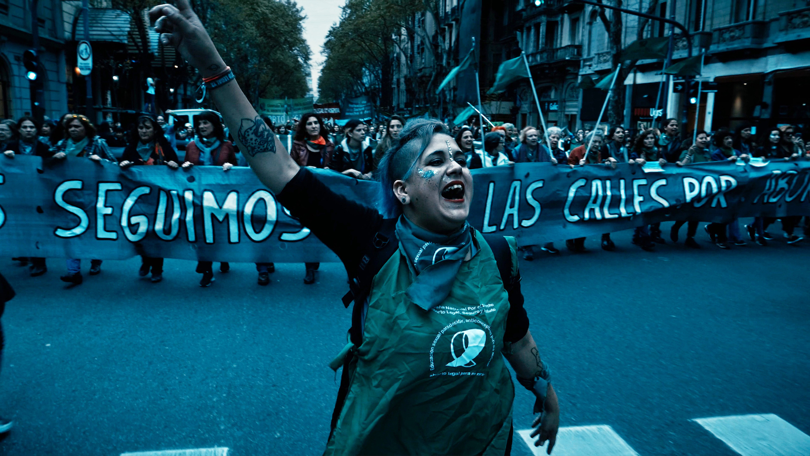 A woman marches in the street in front of a banner, one arm raised in triumph