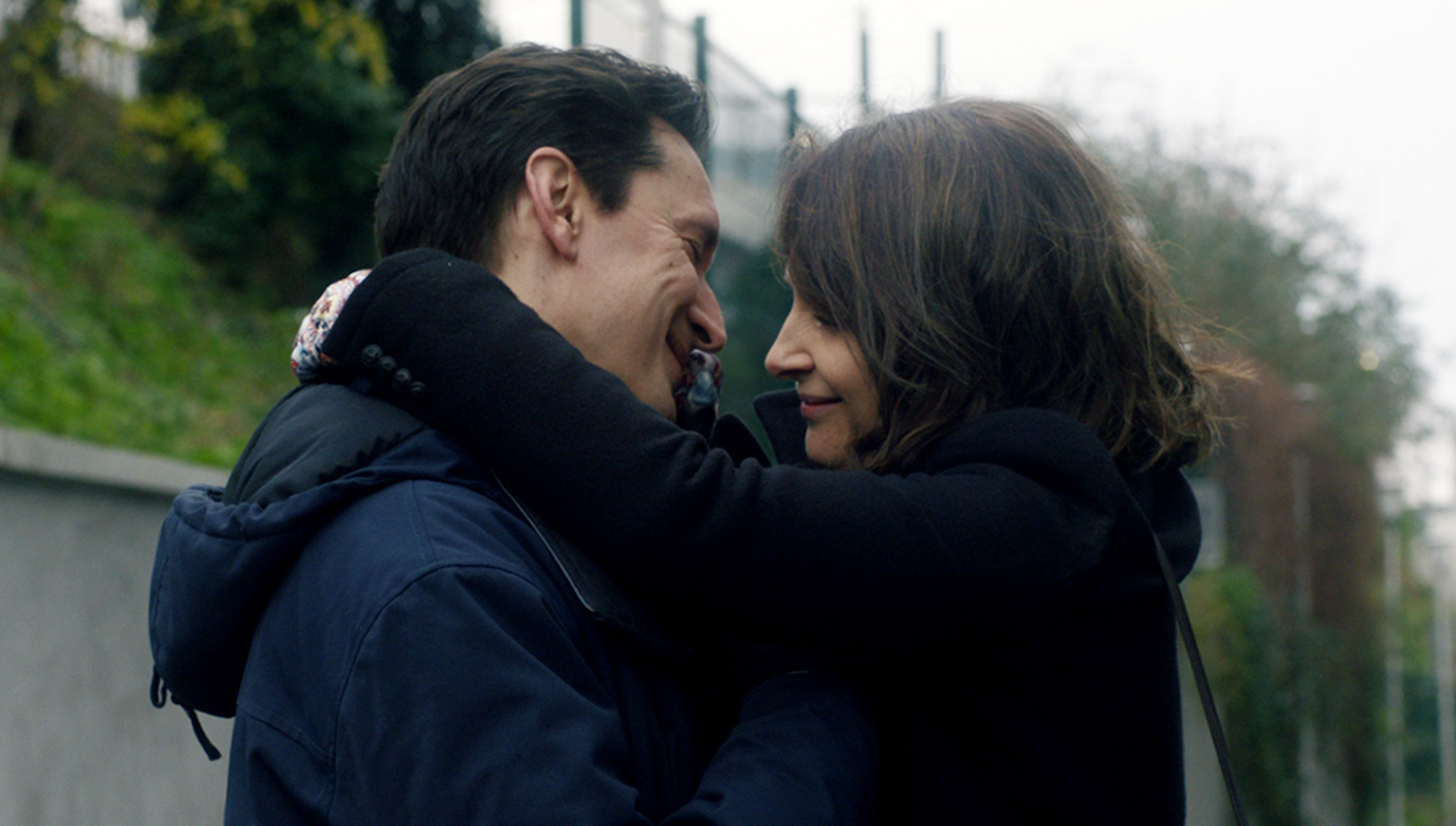 A man and woman hug each other and smile surrounded by green trees