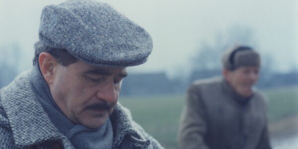 Two men, one in foreground, one blurred in back, stand in a desolate winter landscape.