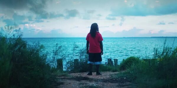 A man stands with back to camera staring out at a blue wide river