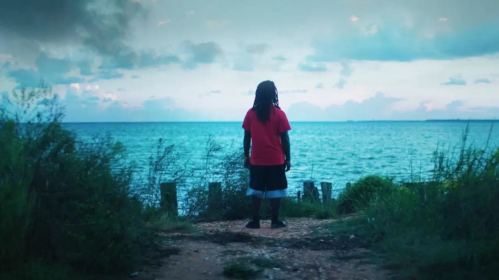 A man stands with back to camera staring out at a blue wide river