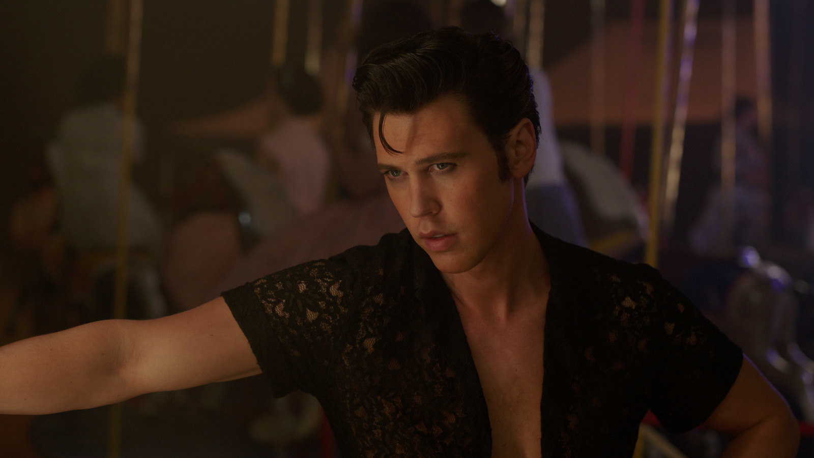 A man in a pompadour and open shirt extends his right arm out and looks offscreen