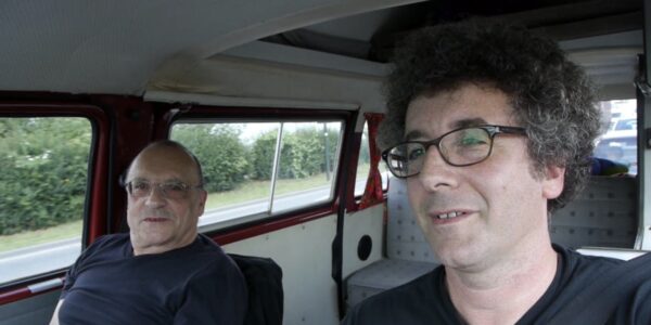 Two men, one older and one younger, sit next to one another in a car, the younger in the driver's seat.