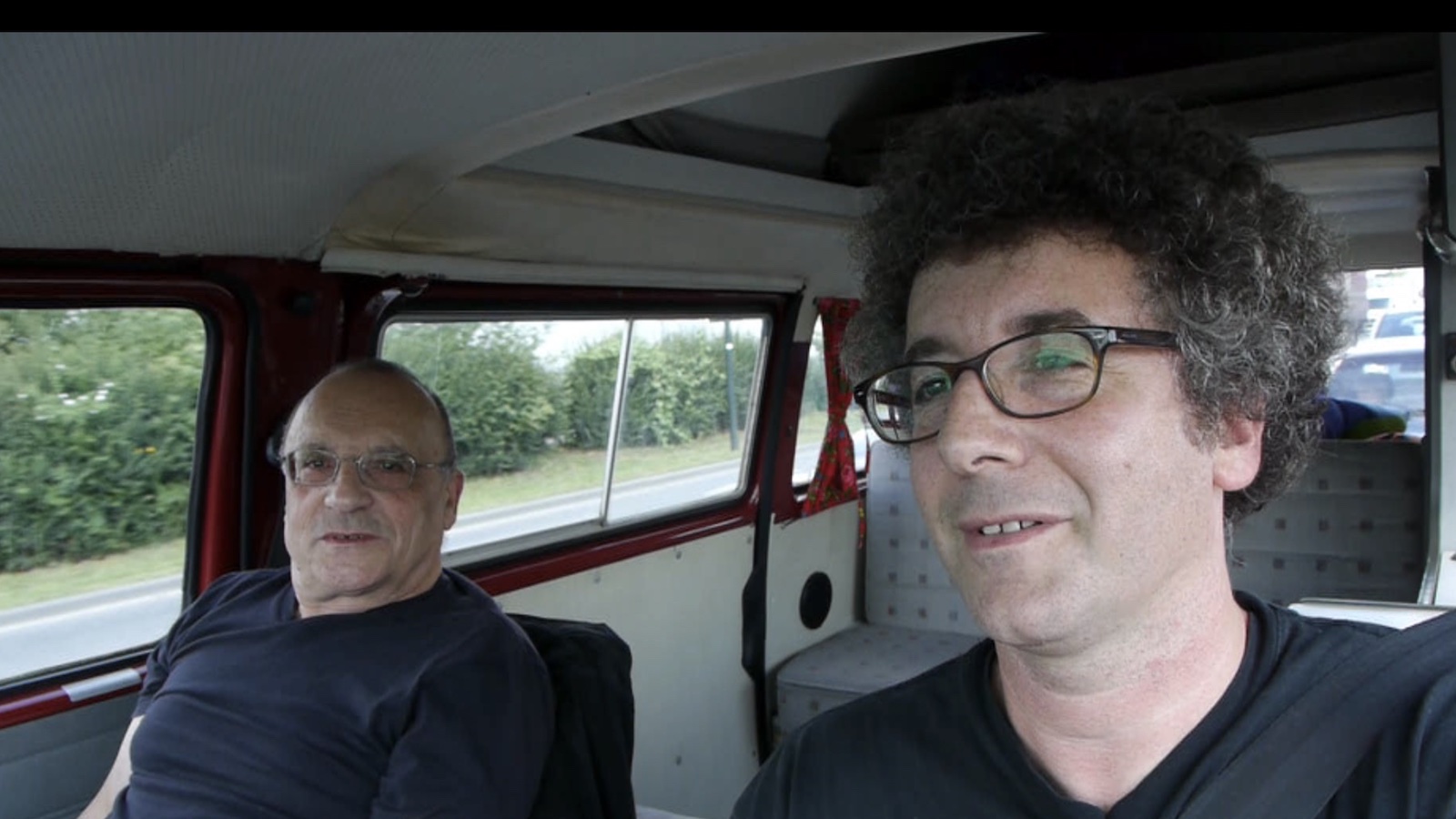 Two men, one older and one younger, sit next to one another in a car, the younger in the driver's seat.