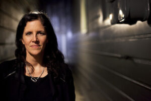 A woman with dark hair stands in a dimly lit hallway and looks at camera half smiling