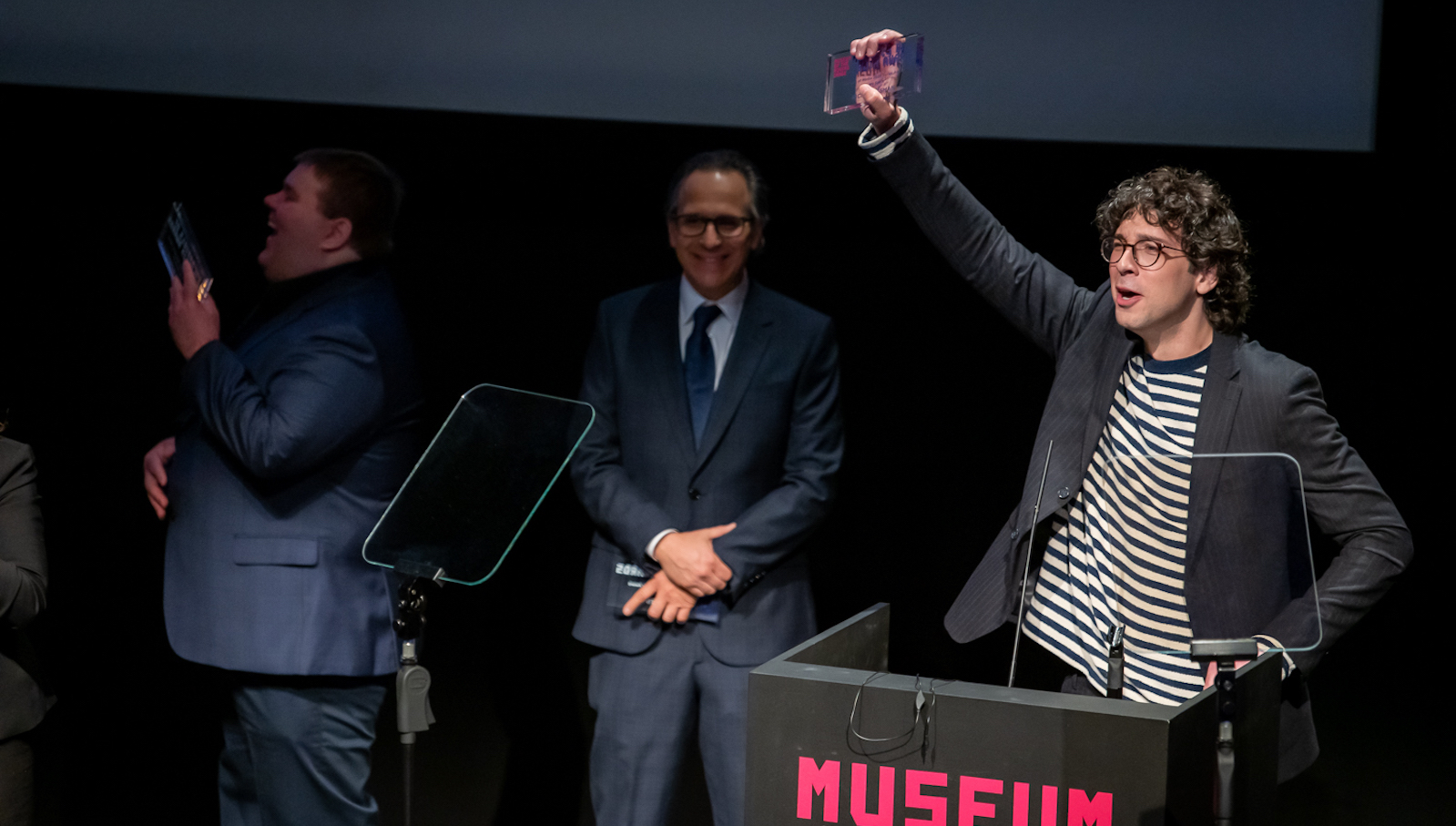A man in striped shirt holds up an award at the Museum stage podium.