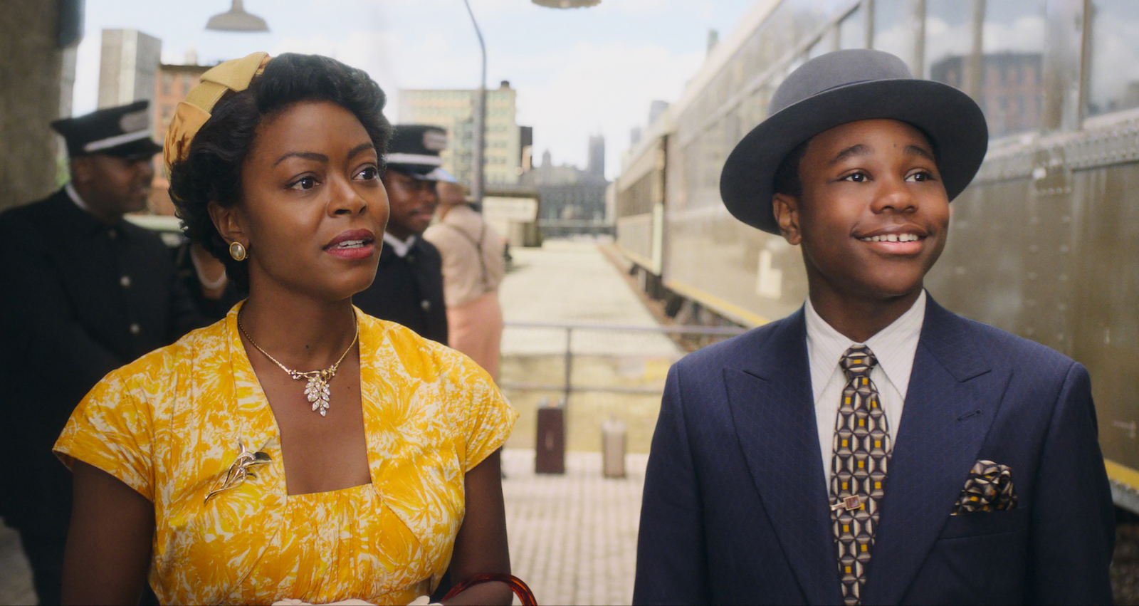 A mother in a yellow dress and her son, in a blue suit and fedora stand on a train platform.