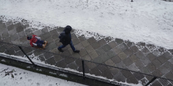 An adult figure pulls a child in a small sled on a wintry street as seen from a high balcony.