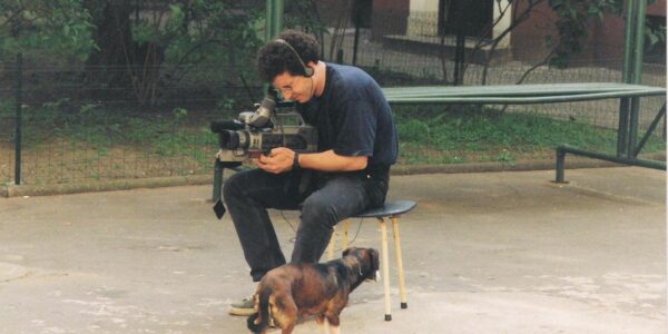 A man sitting in a park looks through a video camera viewfinder while a dog walks by.