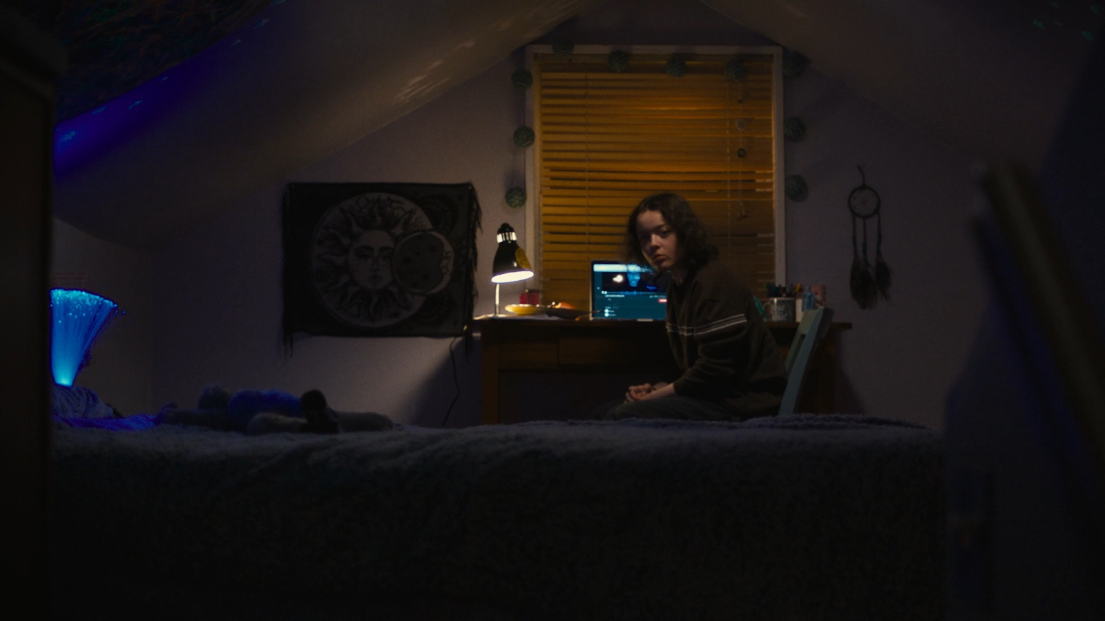 A teenage girl in an attic bedroom looks back at camera from her desk and laptop