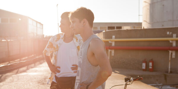 Two young men, the one in the foreground wearing a tanktop, look off-screen against a sun-dappled sky by a train track