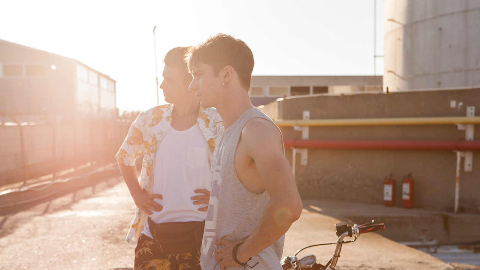 Two young men, the one in the foreground wearing a tanktop, look off-screen against a sun-dappled sky by a train track