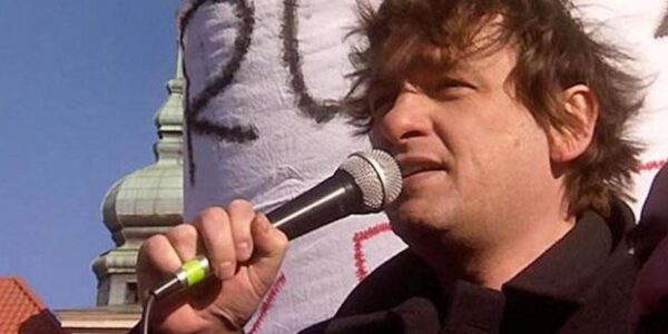 A man speaks into a microphone in front of a handmade sign