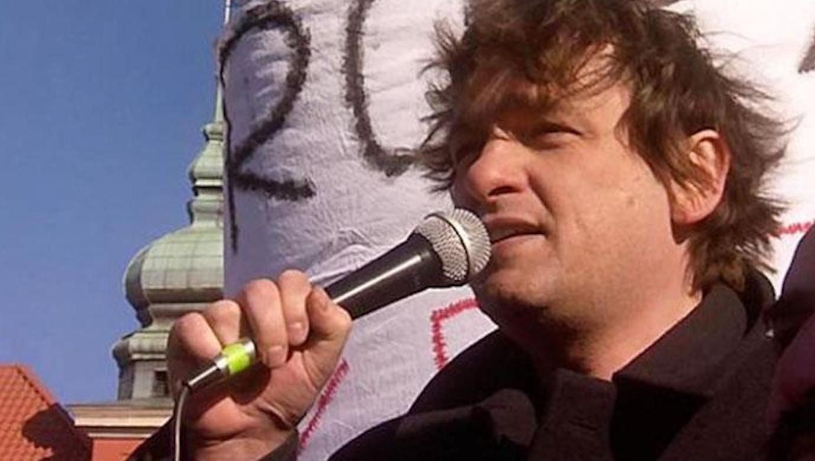 A man speaks into a microphone in front of a handmade sign